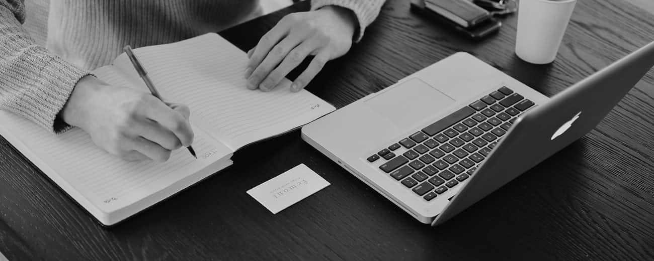 A hand writing in an open notebook, next to a laptop and a cup on a desk.
